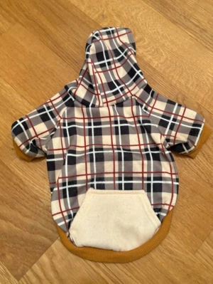 Rutig hundtröja med huva - Säljer en söt hundtröja med huva i rutigt mönster i beige, svart och rött. Tröjan har en praktisk ficka på ryggen och muddar i senapsgult. Perfekt för att hålla din hund varm och snygg under promenader.