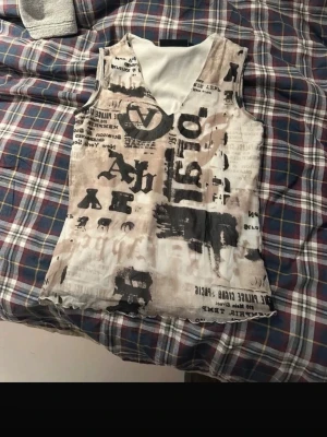 Beige och svart linnetopp från KappAhl - Cool ärmlös linnetopp från KappAhl med beige och svart tryck i graffiti-stil. Toppen har v-ringning och är gjord i ett lätt och mjukt material, perfekt för en avslappnad och trendig look.