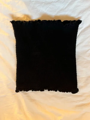 Svart bandeau tubtopp - Säljer en svart bandeau tubtopp med smockad stretch och vågig kant upptill och nedtill. Perfekt att styla till jeans eller kjol för en enkel och trendig look.
