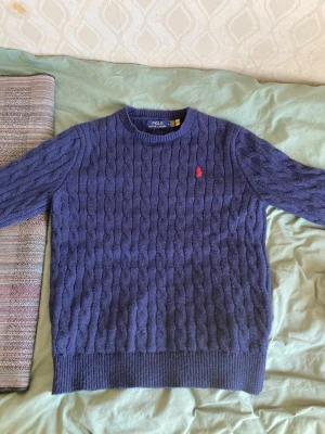 Stickad Ralph lauren tröja - Mörkblå kabelstickad tröja från Polo Ralph Lauren med rund halsringning och röd broderad logga på bröstet. Tröjan har ribbstickade muddar vid ärmslut och nederkant. Perfekt för lager-på-lager och klassisk stil.