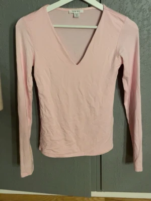 Ljusrosa v-ringad topp från Amisu - Söt ljusrosa långärmad topp från Amisu med djup v-ringning. Tillverkad i mjuk bomullsmix som sitter snyggt och bekvämt. Perfekt att styla med jeans eller kjol för en enkel och fräsch look.