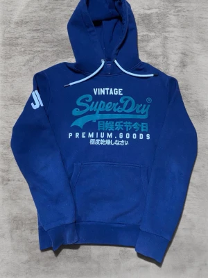 Blå Superdry hoodie storlek L - Snygg blå hoodie från Superdry med stort tryck i turkos och vitt på bröstet samt japanska tecken. Klassisk känguruficka framtill, huva med vita dragsnören och ribbade muddar. Perfekt för en avslappnad streetstyle-look.              Tröjan är i mycket bra skick bortsett lite lös söm i armhålan som man ser på bild 5. Syns inte när man använder tröjan och om man ogillar kan man nog lätt fixa ihop det igen.