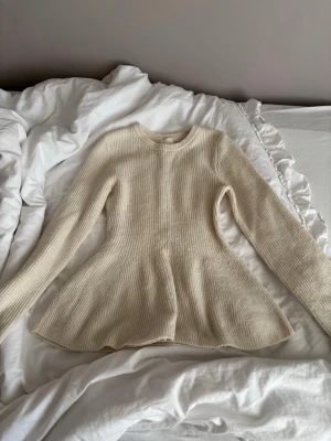 Krämvit peplumstickad tröja - Supersnygg krämvit stickad tröja med peplumformad midja och långa ärmar. Tröjan har rund halsringning och ribbstickad struktur som ger en stilren look. Perfekt för dig som vill ha något både bekvämt och trendigt. Off White färg