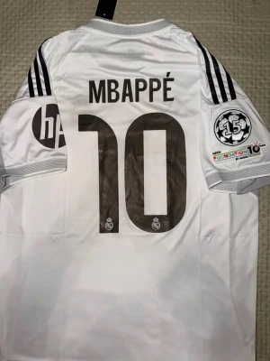 Vit Real Madrid Mbappé tröja - Säljer en vit Real Madrid matchtröja med Mbappé och nummer 10 på ryggen. Tröjan har svarta detaljer, tre ränder på axlarna och flera tryck på ärmarna. Perfekt för fotbollsfans som vill sticka ut på läktaren eller planen.