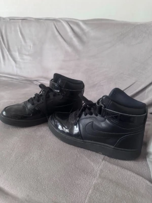 Svarta Nike mid sneakers i skinn - Snygga svarta sneakers från Nike i mid-modell med klassisk swoosh på sidan. Skorna är gjorda i blankt och matt skinn, har svarta skosnören och kardborreband vid ankeln. Perfekta för en streetwear-look.