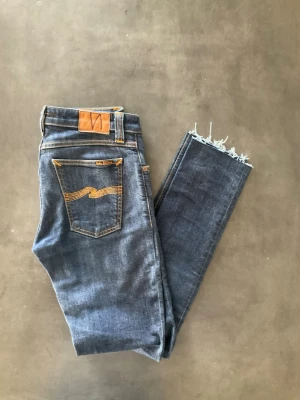 Mörkblå jeans från Nudie Jeans - Snygga mörkblå jeans från Nudie Jeans med klassiska orange sömmar och vågig detalj på bakfickan. Jeansen har rå kant nertill för en trendig look och är tillverkade i denim.  Perfekta för dig som gillar en avslappnad men stilren stil.
