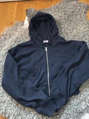 Mörkblå hoodie från Gina Tricot - Mörkblå hoodie från Gina Tricot med dragkedja framtill och två stora fickor. Klassisk huva med dragsko och ribbade muddar vid ärmslut och nederkant. Perfekt för en avslappnad och cool stil. Tillverkad i mjuk bomullsblandning.