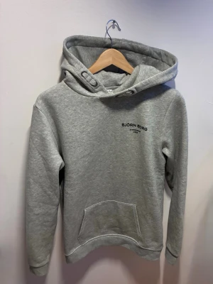 Grå hoodie från Björn Borg - Snygg grå hoodie från Björn Borg med svart logga på bröstet. Klassisk känguruficka framtill och justerbar huva med dragsko. Mjuk och skön bomullsblandning, perfekt för en avslappnad stil. Helt ny utan prislapp 