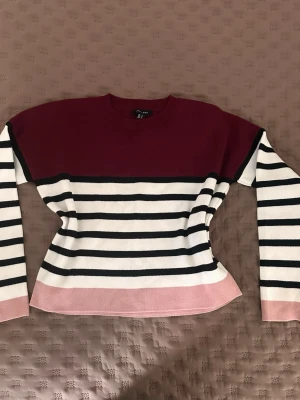 Randig stickad tröja från New Look - Snygg stickad tröja från New Look med vinrött parti upptill, svarta och vita horisontella ränder samt ljusrosa kant nertill och vid ärmsluten. Tröjan har rund halsringning och långa ärmar. Perfekt för dig som gillar färgkombinationer och randigt.