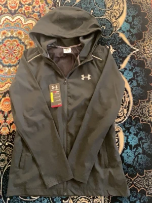 Svart vindjacka från Under Armour - Svart vindjacka från Under Armour med huva och dragkedja framtill. Jackan har vita logotyper på bröstet och ryggen samt diskreta reflexdetaljer vid axlarna. Tillverkad i lätt och vattenavvisande material, perfekt för blåsiga dagar.