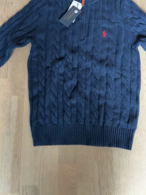 Marinblå kabelstickad tröja Polo - Kabelstickad marinblå tröja från Polo Ralph Lauren med dragkedja vid halsen och röd broderad logga på bröstet. Tröjan har ribbade muddar och är perfekt för lager-på-lager-stil. Klassisk och tidlös design. Hör av er gärna 