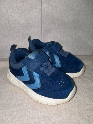 Blå Hummel sneakers med kardborre - Blåa sneakers från Hummel med ljusblå detaljer. Skorna har kardborreband och elastiska skosnören för enkel påtagning. Enda negativa är att plösen lossnat från snörena men inget som man tänker på, se sista bilden.