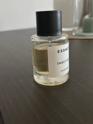 ESSENCE Sweet Cara parfym - Säljer en stilren parfymflaska från ESSENCE med doften Sweet Cara. Flaskan är genomskinlig med svart lock och vit etikett. Innehållet är ljusgult och flaskan har en modern, minimalistisk design. Perfekt för dig som gillar söta dofter.