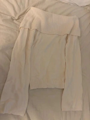 Off shoulder stickad tröja cremevit - Säljer en cremevit stickad off shoulder-tröja med bred ribbad kant upptill och vid ärmslut. Tröjan har lång ärm och är gjord i mjukt stickat material, perfekt för en trendig och avslappnad look.