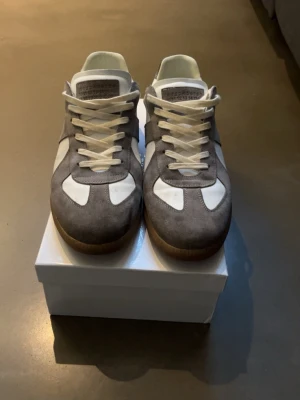 Grå och vita sneakers från Maison Margiela  - Snygga sneakers från Maison Margiela i grå mocka och vitt läder med klassisk gummisula. Skorna har vita snören och en stilren, retroinspirerad design som passar perfekt till streetwear. Tungan har diskret branding och insidan är fodrad i vitt.