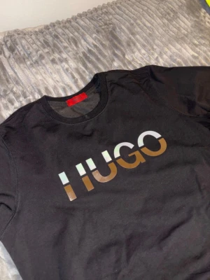 Svart sweatshirt från Hugo - Svart sweatshirt från Hugo med rund halsringning och stort tryckt logotyp i metallicfärger på bröstet. Tröjan har långa ärmar och ribbade muddar vid ärmslut och nederkant. Perfekt för en stilren och trendig look.