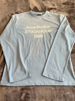 Acne Studios SPRAYED 1996 LOGO T-SHIRT - Snygg ljusblå långärmad tröja från Acne Studios med tryck 'STOCKHOLM 1996' framtill. Tillverkad i mjuk bomull och har en avslappnad passform. Perfekt för dig som gillar minimalistisk och stilren design.