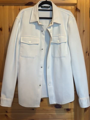 Zara Overshirt  - Stilren vit overshirt från Zara med klassisk krage, tryckknappar och två bröstfickor med lock. Overshirten har långa ärmar med manschett och är tillverkad i ett mjukt tyg som ger en clean och modern look. Pris kan alltid diskuteras💫💫