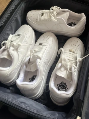 Nike Air Force 1 vita sneakers - Klassiska Nike Air Force 1 sneakers i helvitt läder med perforerad tå, vit sula och ikonisk Swoosh på sidan. Skorna har låga skaft och metall-detalj på snörningen. Perfekta för en clean och stilren look.