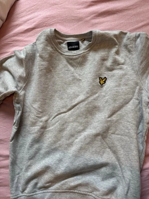 Grå sweatshirt från Lyle & Scott - Snygg grå sweatshirt från Lyle & Scott med klassisk rund halsringning och gul logga broderad på bröstet. Tröjan har ribbade muddar vid ärmslut och nederkant. Perfekt för en avslappnad stil.