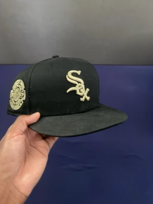 Svart New Era White Sox keps - Säljer en svart New Era 59FIFTY keps med White Sox-logga broderad i vitt framtill och ett detaljerat sidomärke. Klassisk flat brim och strukturerad passform. Perfekt för dig som gillar sportig streetstyle och vill ha en ikonisk look.