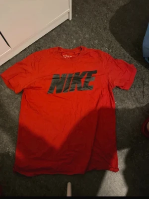 Röd Nike t-shirt med stor logga - Röd t-shirt från Nike med stor svart logga över bröstet. Klassisk rund halsringning och korta ärmar. Tillverkad i mjuk bomull som är skön att ha på sig. Perfekt för en sportig och avslappnad stil.