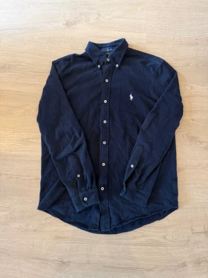 Mörkblå skjorta från Ralph Lauren - Klassisk mörkblå skjorta från Ralph Lauren med button-down krage och broderad logga på bröstet. Skjortan har långa ärmar, vita knappar och är tillverkad i mjuk bomull. Perfekt för en stilren look.