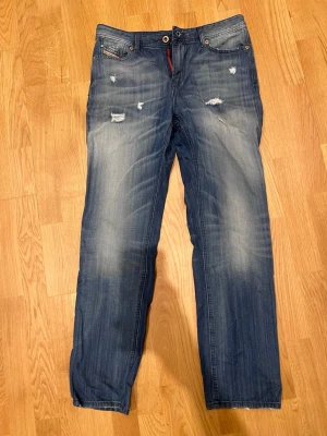 Blå slitna jeans från Diesel - Säljer ett par blå jeans från Diesel, modell Reen, med regular-straight passform och normal midja. Jeansen har slitna detaljer och ljusare partier framtill för en cool, avslappnad look. Klassisk femficksmodell och knappgylf.
