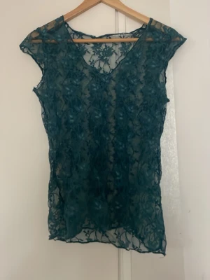 Grön spetsblus med v-ringning - Snygg grön blus i transparent spets med v-ringning och korta ärmar. Blusen har ett blommigt mönster i spetsen och är perfekt för att skapa en trendig lager-på-lager-look.