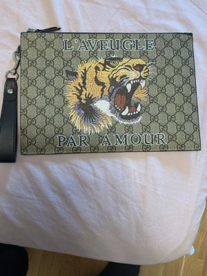 Hand  kuvertväska med tigertryck - Kuvertväska  i beige canvas med klassiskt GG-mönster, broderad tiger och texten 'L'Aveugle Par Amour' på framsidan. Svart avtagbart handledsband i läder och dragkedja upptill. Perfekt för att sticka ut med en lyxig touch.