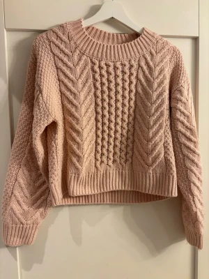 Ljusrosa kabelstickad tröja - Mysig ljusrosa stickad tröja med grovt kabelmönster framtill och på ärmarna. Tröjan har rund halsringning, ribbade muddar och en något boxig passform. Perfekt för lager-på-lager och chill dagar.