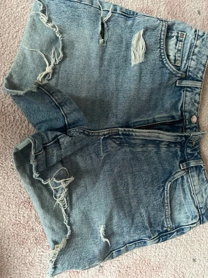 Blå slitna jeansshorts  - Säljer ett par blå jeansshorts från Perfect Jeans i storlek 38. Shortsen har en hög midja, råa kanter och slitna detaljer framtill för en trendig look. Klassisk femficksmodell med dragkedja och knapp. Perfekta till sommaren!