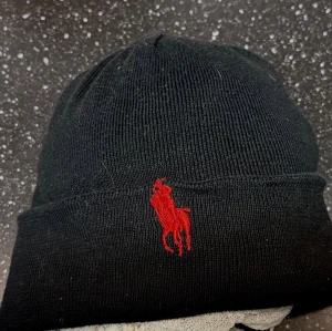 Svart mössa från Polo Ralph Lauren - Svart ribbstickad mössa från Polo Ralph Lauren med klassisk röd broderad logga framtill. Enkel och stilren design med uppvikt kant, perfekt för kalla dagar. Materialet är mjukt och stretchigt för skön passform.