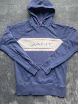 Blå hoodie från GANT - Snygg blå hoodie från GANT med vit panel och röda ränder över bröstet samt stor GANT-logga. Klassisk huva och känguruficka framtill. Perfekt för en avslappnad och stilren look.