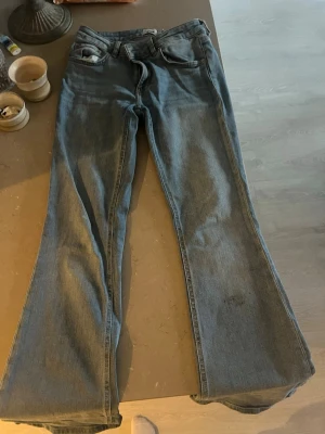 Blå bootcut jeans med låg midja - Säljer ett par klassiska blå bootcut jeans med låg midja och fem fickor. Jeansen har en lätt utsvängd passform nertill och är tillverkade i denim. Perfekta för dig som gillar en retrostil och vill ha ett par snygga vardagsjeans.