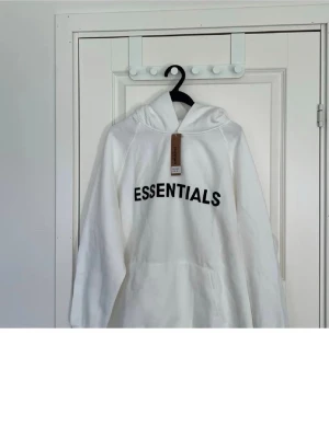 Vit Essentials hoodie med tryck - Snygg vit hoodie från Essentials med svart tryck på bröstet och liten logga på huvan. Klassisk känguruficka framtill och mjukt material som känns skönt mot huden. Perfekt för en avslappnad streetwear-look.