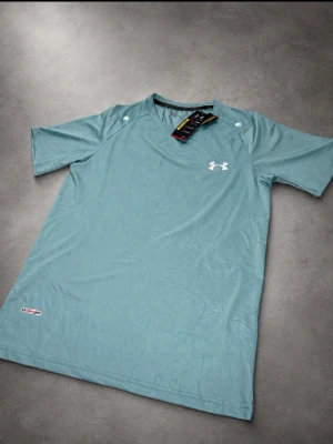 Blågrön sport-t-shirt från Under Armour - Sportig blågrön t-shirt från Under Armour med korta ärmar och rund hals. Tillverkad i ett lätt och snabbtorkande funktionsmaterial, perfekt för träning. Diskret logga på bröstet och liten etikett nertill. Snygg och bekväm för aktiva dagar.