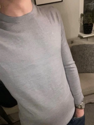 Ljusgrå stickad tröja J.Lindeberg - Snygg ljusgrå stickad tröja från J.Lindeberg med diskret logga på bröstet. Tröjan har rund halsringning, långa ärmar och ribbade muddar vid ärmslut och nederkant. Perfekt till jeans eller chinos för en clean look.