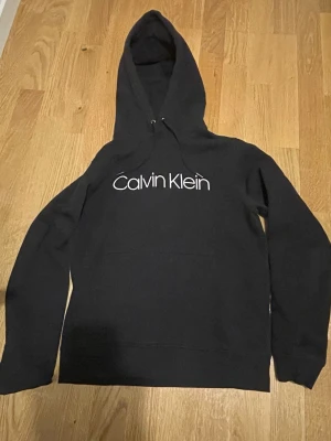 Svart Calvin Klein hoodie XS - Snygg svart hoodie från Calvin Klein med stor vit logga på bröstet. Klassisk modell med huva och dragsnören. Tillverkad i mjukt material som känns skönt mot huden. Perfekt för en avslappnad och trendig look.