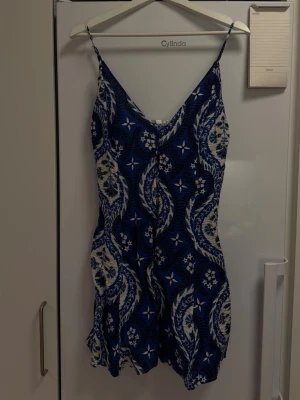 Blåvit kortklänning med mönster - Säljer en blåvit kortklänning med smala axelband och v-ringning. Klänningen har ett blommigt och paisley-inspirerat mönster samt knytning i ryggen. Perfekt för varma dagar och har en luftig passform.