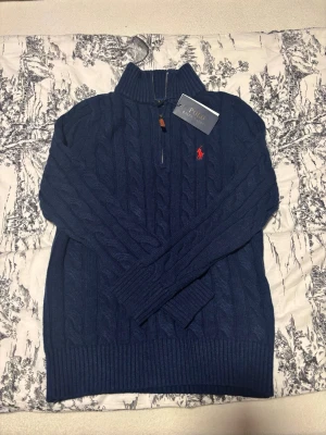 Mörkblå kabelstickad tröja Polo Ralph Lauren - Kabelstickad mörkblå tröja från Polo Ralph Lauren med halv dragkedja och hög krage. Tröjan har det klassiska röda Polo-logot broderat på bröstet och ribbade muddar vid ärmslut och nederkant. Perfekt för en stilren och avslappnad look.