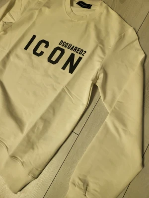 Beige sweatshirt från Dsquared2 ICON - Snygg beige sweatshirt från Dsquared2 med stor svart 'ICON'-logga framtill. Tröjan har rund halsringning, långa ärmar och ribbade muddar vid ärmslut och nederkant. Tillverkad i mjukt bomullsmaterial som känns skönt mot huden. Perfekt för en trendig och avslappnad stil. Och riktig bra kvalitet de lovar jag er 