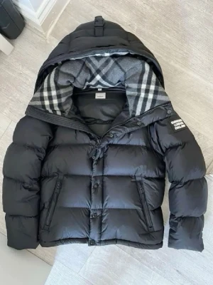 Svart dunjacka från Burberry - OBS DET FINNS ETT LITET HÅL I VÄNSTRA FICKAN Snygg svart dunjacka från Burberry med klassiskt rutigt foder i huvan. Jackan har dragkedja och knappar framtill, två fickor med dragkedja och en patch med Burberry-logga på ärmen. Perfekt för kalla vinterdagar och riktigt stilren look.