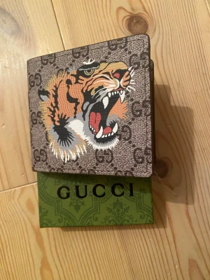 Gucci plånbok med tigertryck - Snygg brun plånbok från Gucci med GG-monogram och ett färgstarkt tigertryck på framsidan. Plånboken har flera kortfack, ett fönster för ID och är gjord i tåligt läder. Perfekt för dig som vill ha något unikt och trendigt i fickan. Tveka inte vid fråger!