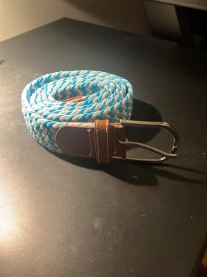 Blåvitt flätat bälte med läderdetaljer - Snyggt flätat bälte i blått och vitt med bruna läderdetaljer och silverfärgat spänne. Perfekt accessoar för att lyfta din outfit med en fräsch färgklick och trendig look. Storlek 105