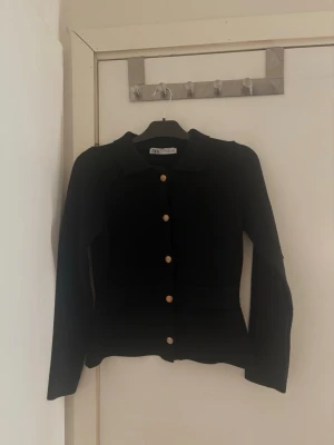 Svart kofta med guldknappar från Zara - Stilren svart kofta från Zara med långa ärmar och klassisk krage. Koftan har fem dekorativa guldknappar framtill och är tillverkad i ett mjukt, stickat material som ger en snygg och bekväm passform. Perfekt att matcha med jeans eller kjol. Storkek S/36/8
