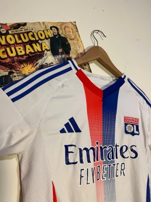 Olympique Lyonnais matchställ Adidas - Säljer ett Olympique Lyonnais matchställ från Adidas. Tröjan är vit med blå och röda detaljer, klubbmärke på bröstet och 'Emirates Fly Better' tryckt framtill. Tillhörande blå shorts med klubbmärke och vita detaljer. Perfekt för fotbollsträning eller match.