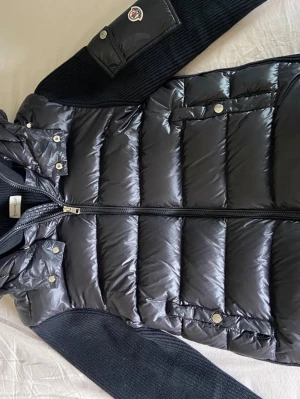 Svart dunjacka från Moncler Size M - Snygg svart dunjacka från Moncler med glansig quiltad kropp och stickade ärmar. Jackan har huva, dragkedja framtill och fickor med tryckknappar. Moncler-logga på ärmen och en extra ficka på armen ger en cool detalj.