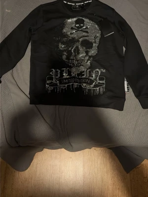 Svart sweatshirt Philipp Plein döskalle - Cool svart sweatshirt från Philipp Plein med glittrande döskalle- och korslagda ben-motiv framtill. Limited Edition-tryck under motivet. Tröjan har rund hals och långa ärmar, perfekt för dig som gillar edgy streetwear.