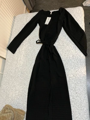 Svart långärmad klänning  - Snygg svart långärmad klänning med knytband i midjan och diskret rutmönster i tyget. Köpt från bubbleroom, prislapp kvar, ALDRIG ANVÄND! Värde 600 kronor